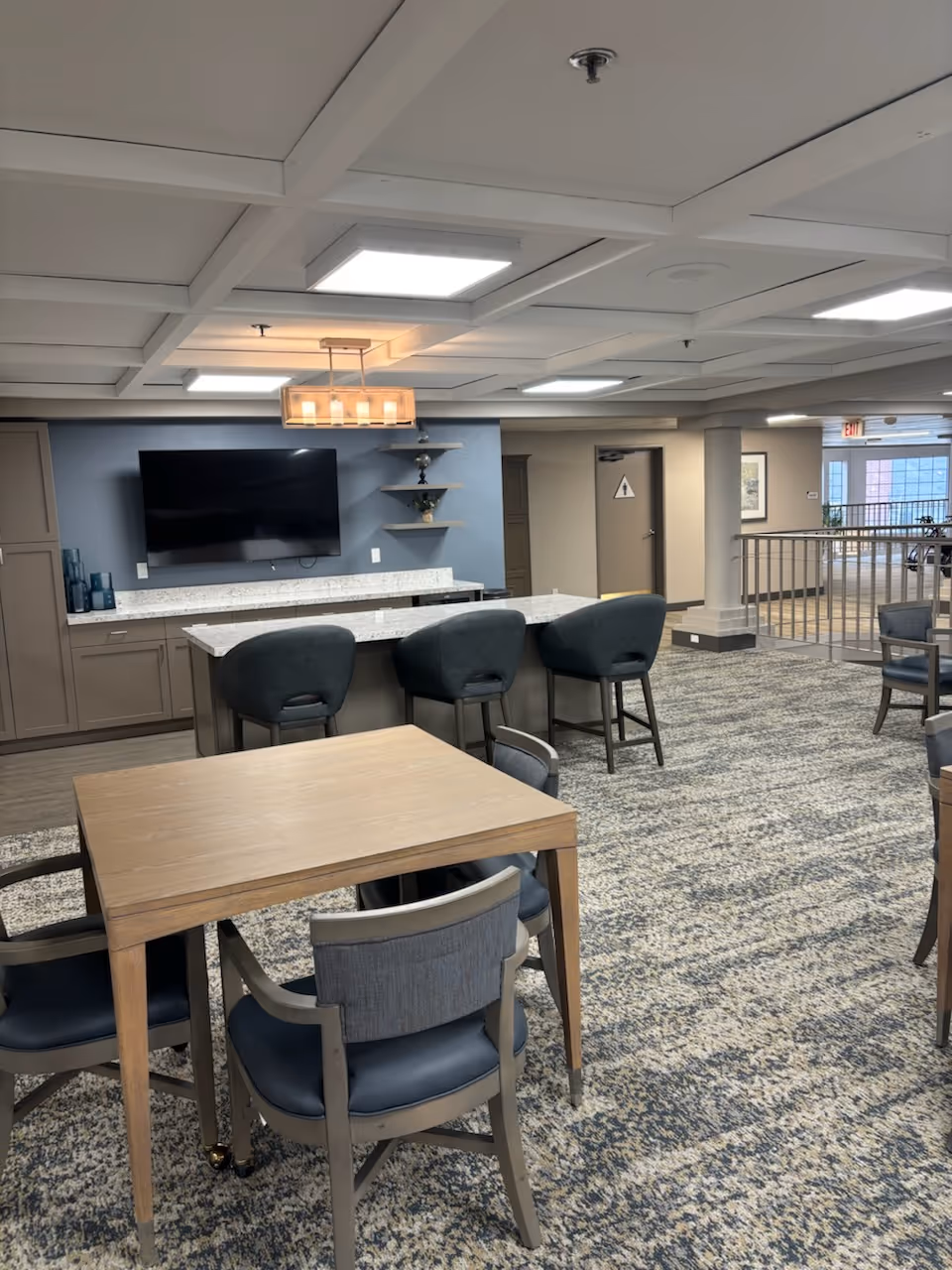 Communal dining and lounge area with tables and chairs, a countertop with bar stools, and a wall-mounted TV.