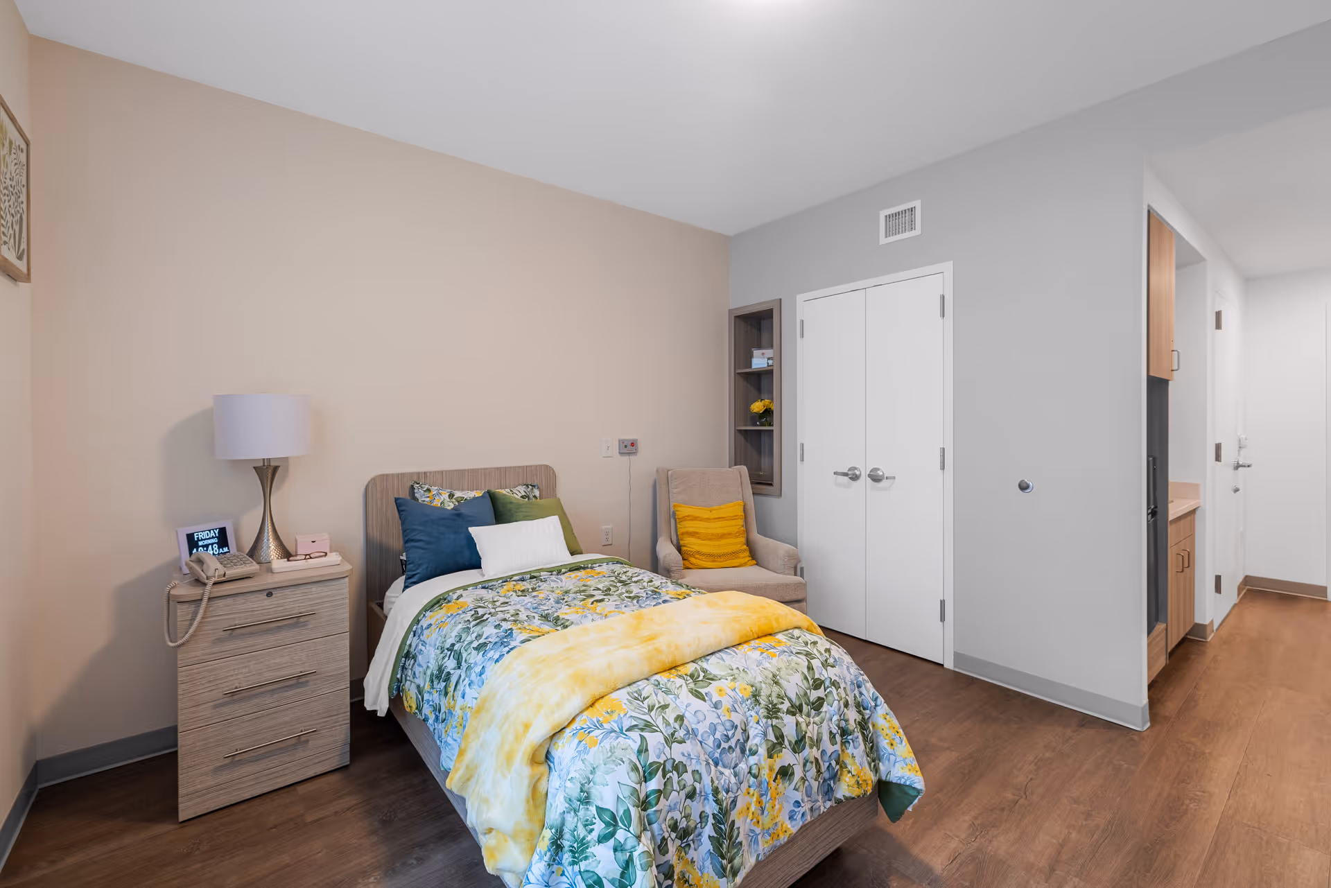 A cozy bedroom in a senior living facility featuring a single bed with floral and yellow bedding, a wooden nightstand with a lamp, phone, and digital clock, a beige armchair with a yellow cushion, and a closet with double doors. The room has wood flooring and neutral-colored walls.