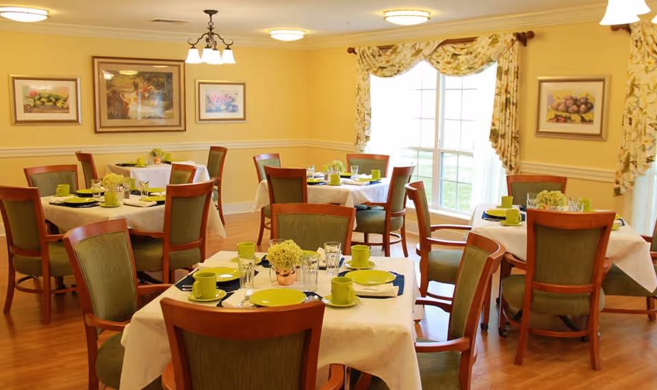 A bright dining room with several round tables covered with white tablecloths, each set with green plates, cups, and glasses. Wooden chairs with green upholstery surround the tables. The room has yellow walls adorned with framed artwork and large windows with floral curtains letting in natural light.