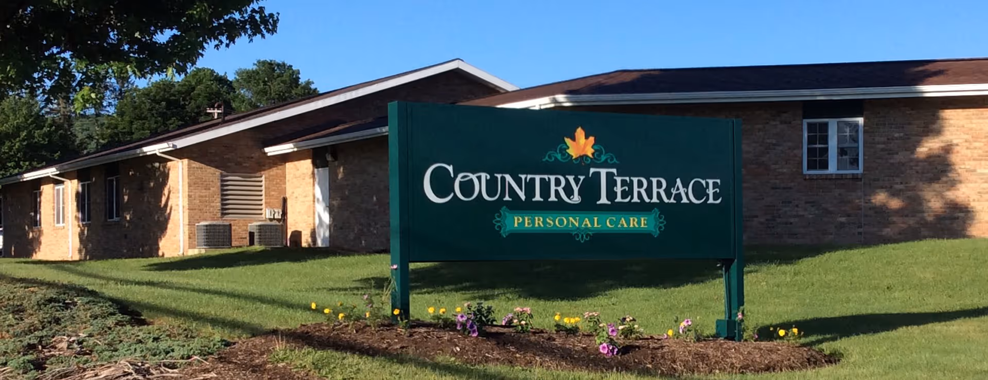 Exterior view of a single-story brick building with a green sign in front that reads 'Country Terrace Personal Care'. The sign is surrounded by a small flower bed with purple and yellow flowers, and there are trees and a clear blue sky in the background.