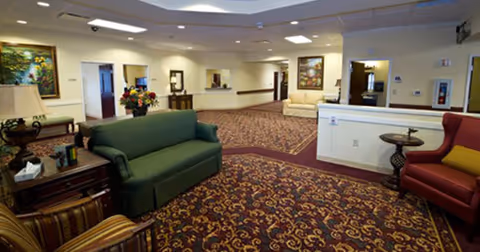 A spacious senior living facility common area with patterned carpet, green and red upholstered chairs and sofas, side tables with lamps and flowers, and framed artwork on the walls. The room is well-lit with ceiling lights and has multiple doorways leading to other rooms.