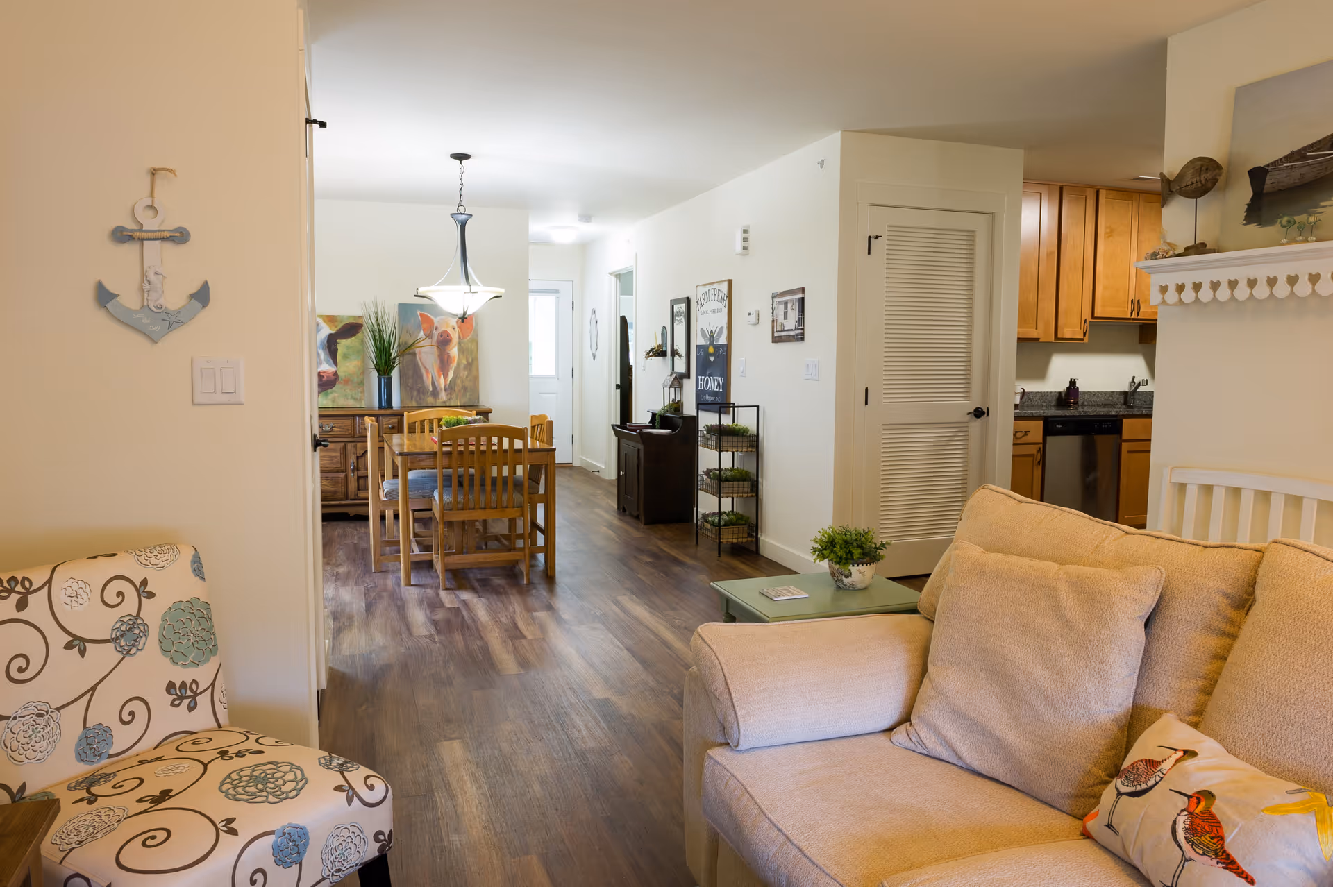 Open-plan interior showing a beige sofa and patterned armchair in the foreground, a wooden dining table and chairs in the middle, and a kitchen area to the right.