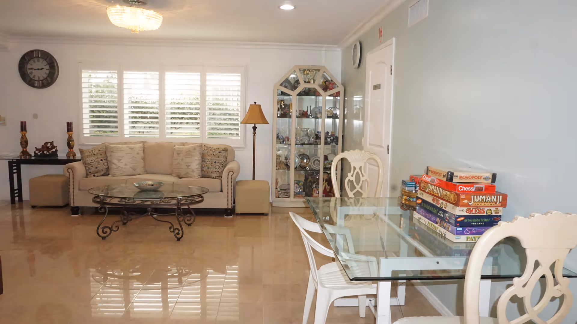 A bright and clean living room area with a beige sofa adorned with patterned cushions, a glass coffee table with ornate metal legs, and a glass display cabinet filled with decorative items. To the right, there is a glass table with white chairs and a stack of board games including Monopoly, Chess, and Jumanji. The room has light-colored walls, a clock on the wall, and a floor lamp next to the sofa.