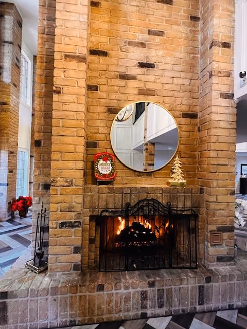 A cozy brick fireplace with a black metal screen in front of a burning fire. Above the fireplace is a round mirror reflecting part of the room. On the mantel are a small decorative Christmas tree and a red holiday-themed sign. The surrounding area features brick walls and a patterned floor with a glimpse of other interior spaces.