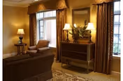 A cozy living room area with a brown sofa, an armchair, a side table with a lamp, a decorative chest with two lamps and a mirror above it, and large windows with brown curtains allowing natural light to enter.