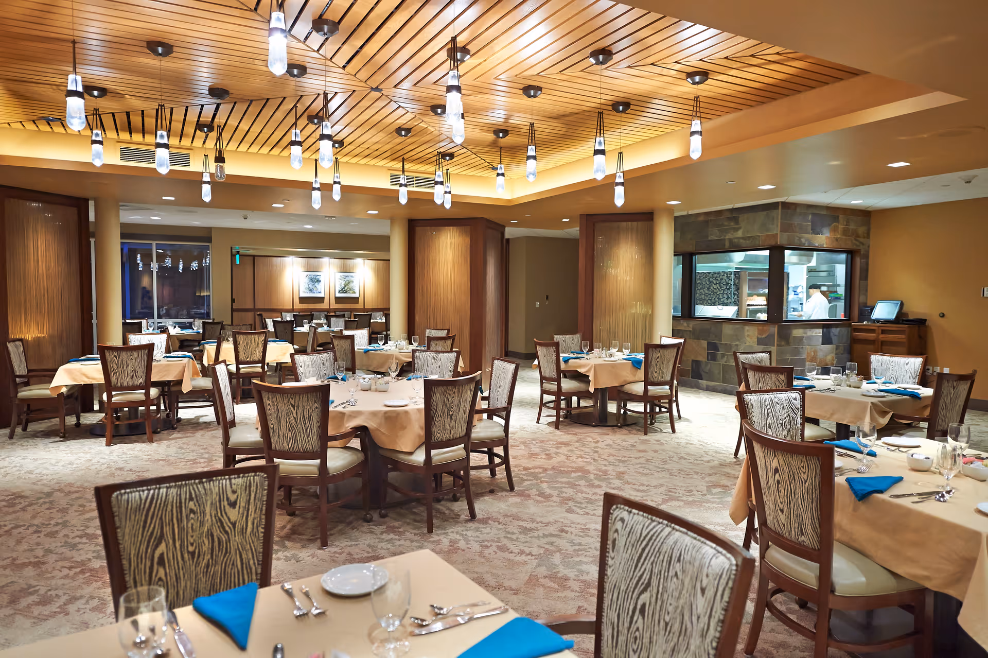 A spacious dining room with multiple tables covered in beige tablecloths and set with plates, glasses, silverware, and blue napkins. The room features wooden chairs with patterned upholstery, a wooden ceiling with modern hanging light fixtures, and a windowed kitchen area where a chef is visible.