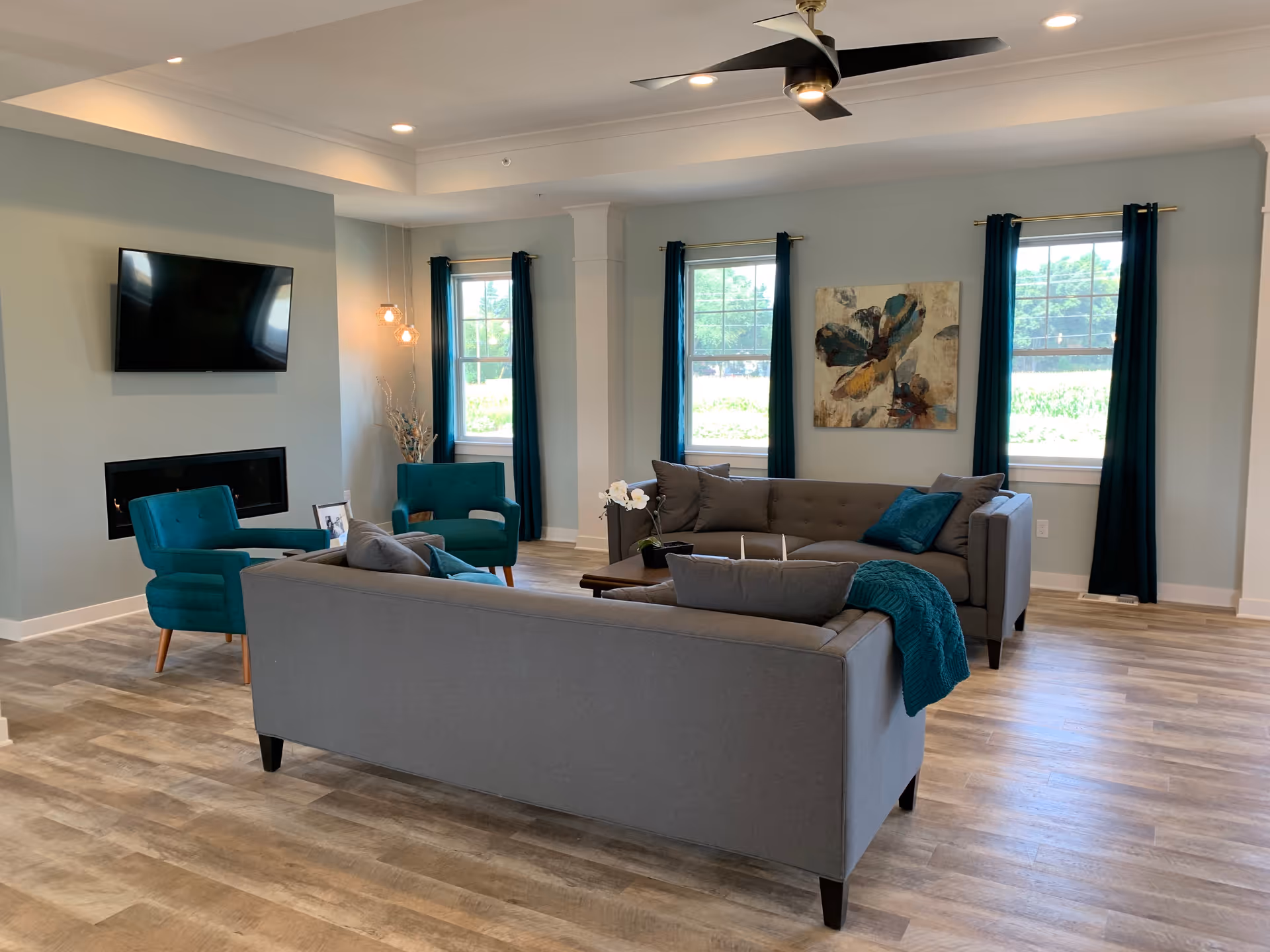 Spacious furnished living room with gray sofas, teal armchairs, wall-mounted TV, fireplace, and windows with dark curtains.