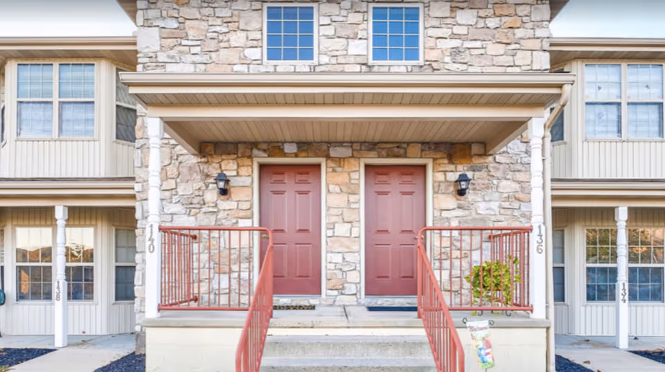 Front exterior view of a senior living facility showing two adjacent red doors with stone and beige siding walls, each door having a small porch with red railings and steps leading up. The building has multiple windows and white pillars supporting the porch roof.
