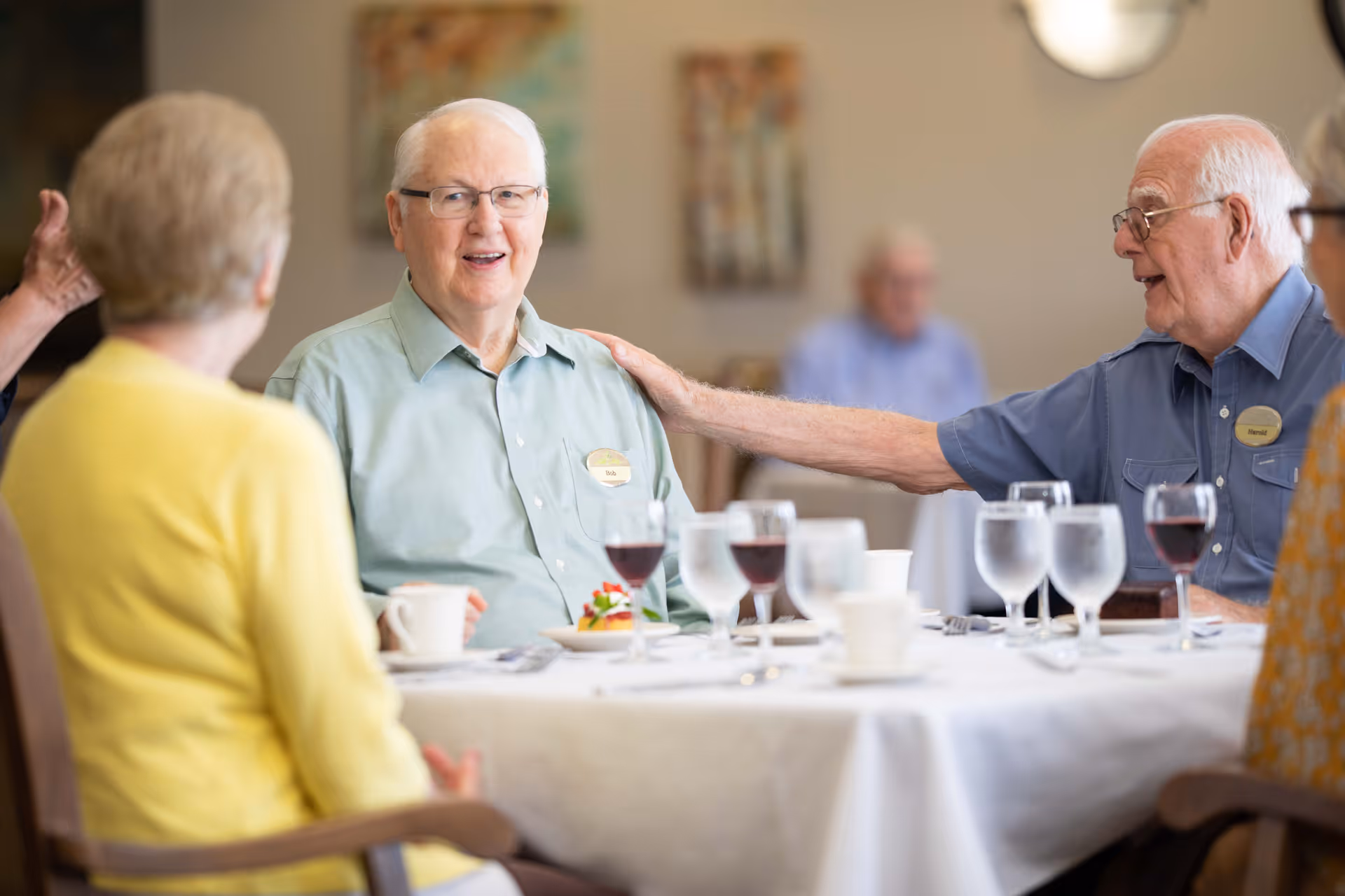 A group of elderly people sitting around a dining table in a senior living facility, engaging in conversation. The table is set with glasses of water, wine, coffee cups, and a dessert plate. One man is smiling and another is reaching out to touch his shoulder in a friendly gesture.