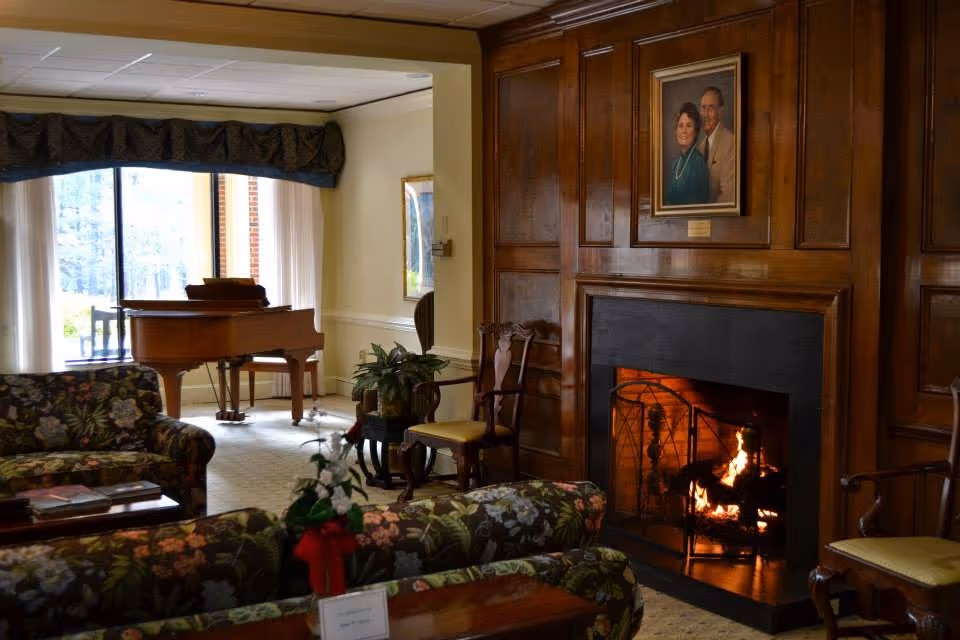 Cozy common room with a lit fireplace set in wood paneling, floral sofas, chairs, and a grand piano by large windows.
