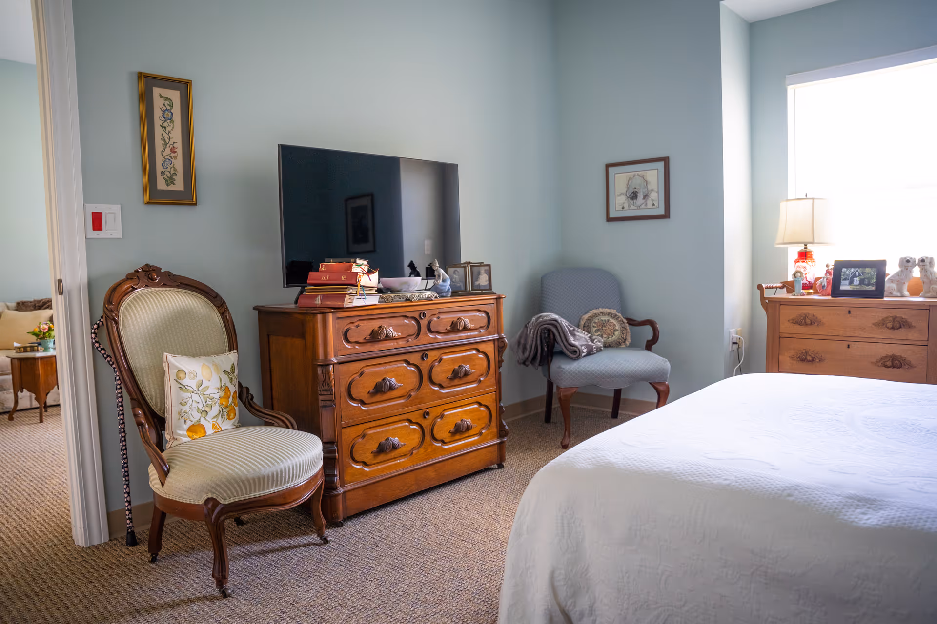 A cozy bedroom with light blue walls featuring a wooden dresser with a flat-screen TV on top, an upholstered armchair with a blanket and pillow, another vintage-style chair with a floral cushion, a bed with a white quilt, and a window letting in natural light. There are framed pictures on the walls and on the dresser.