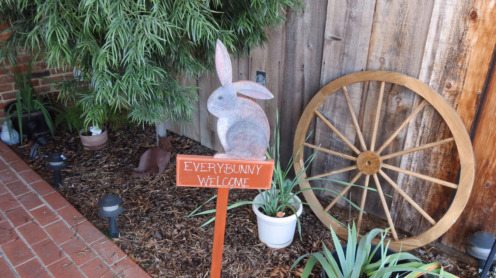 A garden area with a wooden fence backdrop, featuring a decorative wooden rabbit sign that reads 'EVERYBUNNY WELCOME', a large wooden wagon wheel leaning against the fence, several potted plants, and small garden lights along a brick pathway.