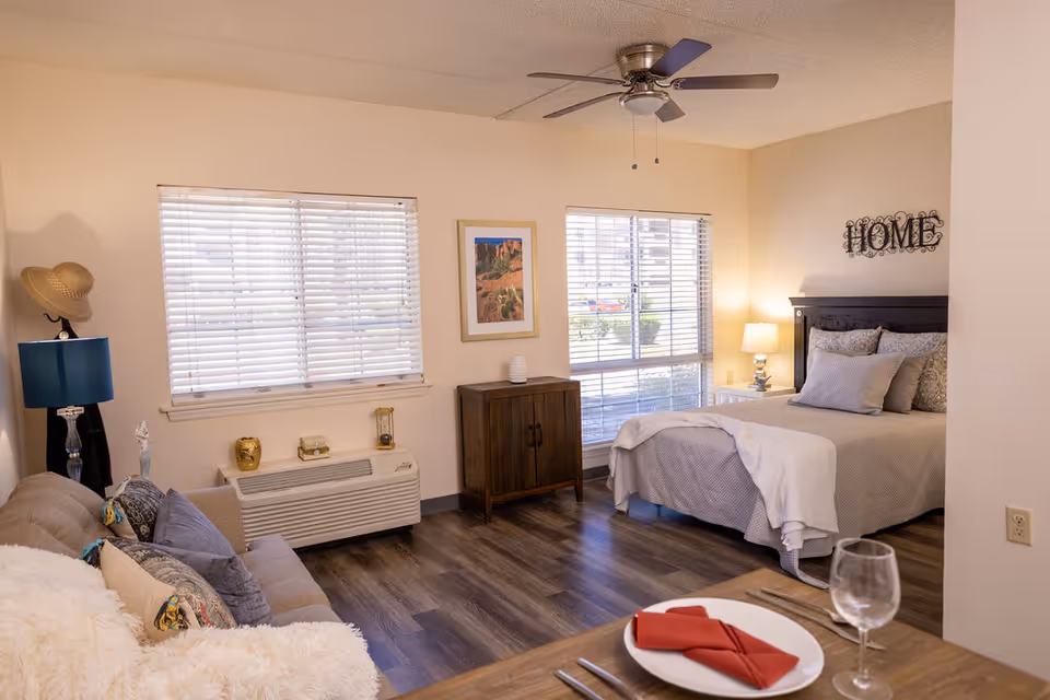A cozy studio apartment with a bed, a small wooden cabinet, a couch with pillows, a lamp, and two windows with blinds letting in natural light. A ceiling fan is mounted above, and a dining table with a plate, red napkin, and wine glass is partially visible in the foreground. The wall above the bed has a decorative 'HOME' sign.