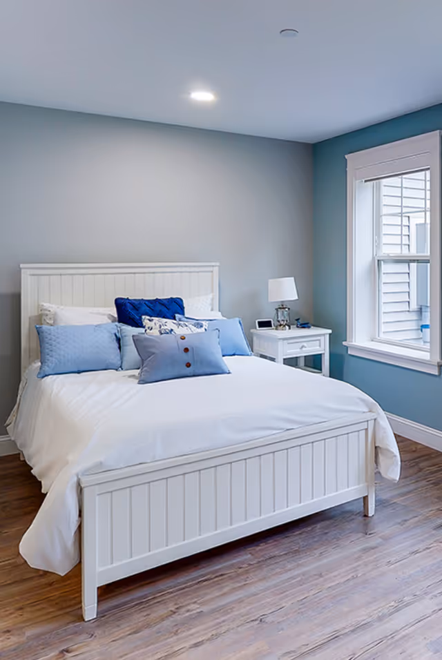 A neatly made bed with white frame and white bedding, adorned with multiple blue and patterned pillows. Next to the bed is a white nightstand with a lamp, a small clock, and some items on it. The room has light blue walls, a window with white trim, and wood flooring.