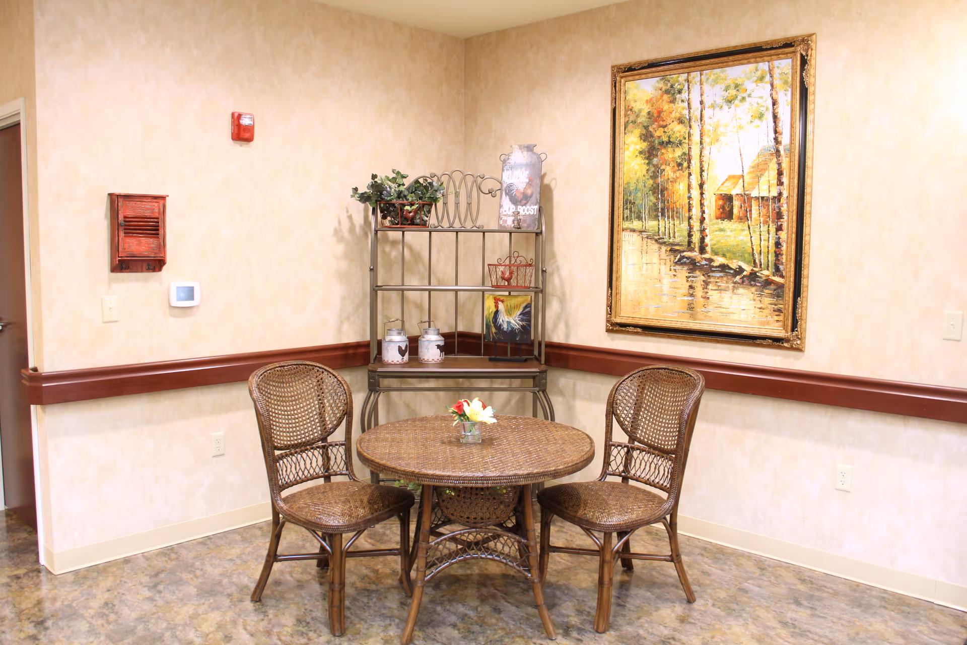 Small seating area with a round wicker table and three wicker chairs beside a decorative shelving unit and a framed landscape painting on the wall.