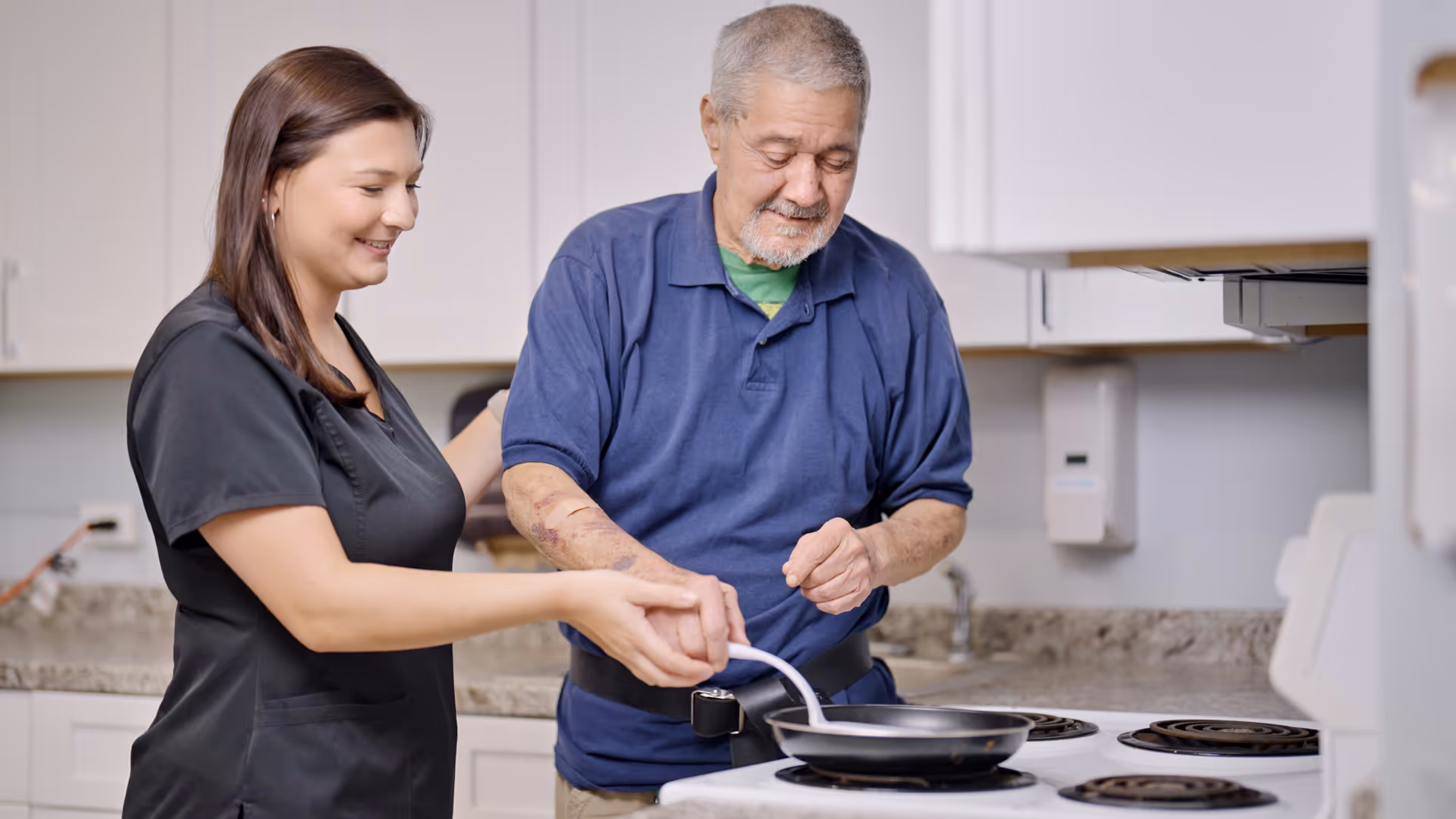 A female caregiver assists an elderly man in cooking on a stove in a kitchen setting. The caregiver is smiling and guiding the man's hand as he holds a spatula over a frying pan. The background shows kitchen cabinets and a countertop.