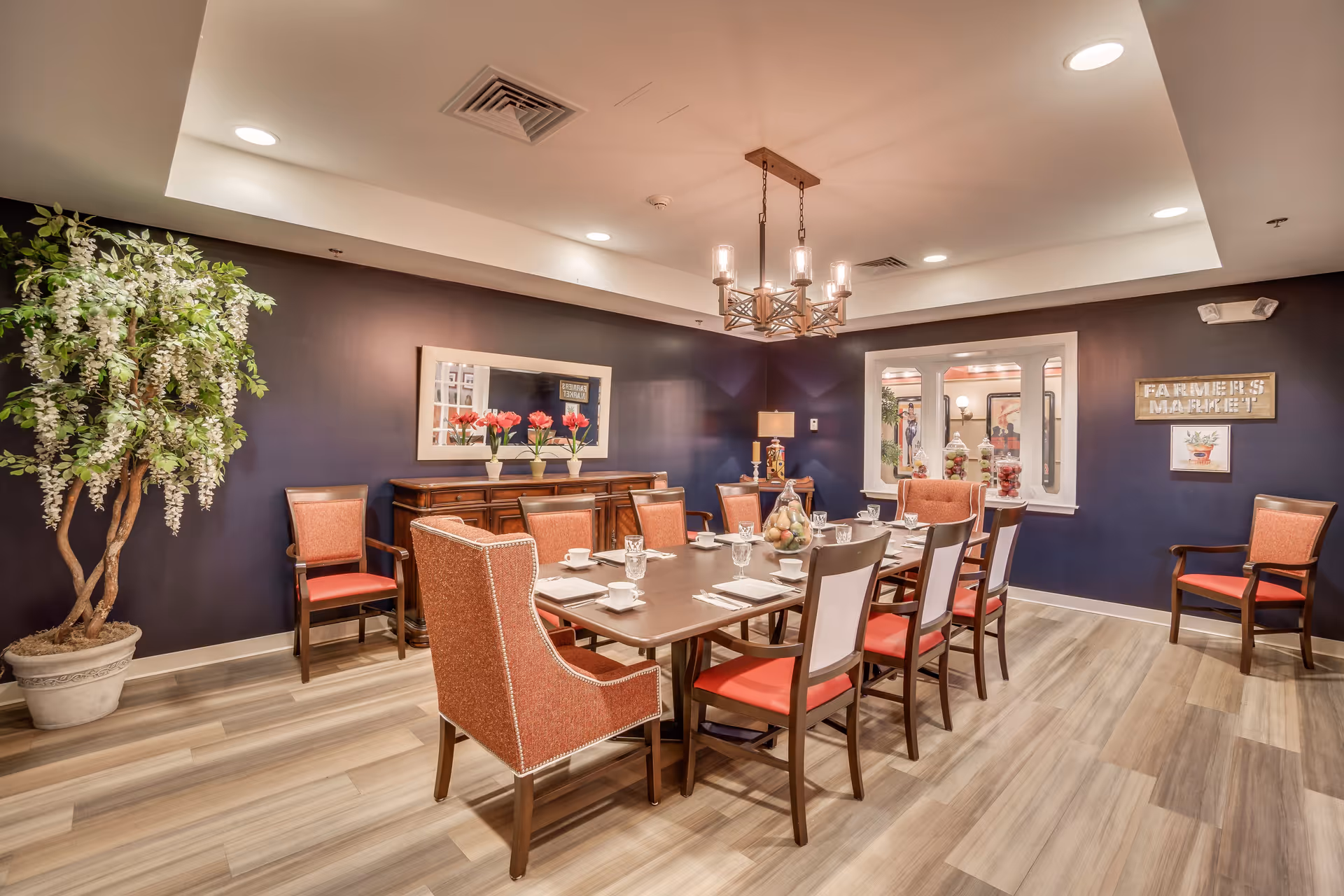 A well-lit dining room with a long wooden table set for a meal, surrounded by chairs with orange upholstery. The room has dark blue walls, a large potted plant in the corner, a sideboard with flower vases, a chandelier hanging from the ceiling, and a window looking into another room. There are decorative signs on the walls, including one that says 'FARMERS MARKET'.