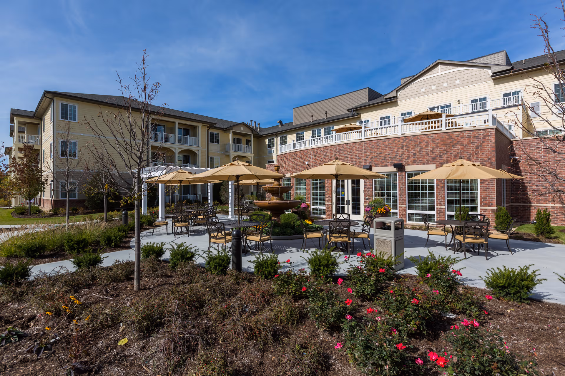Outdoor patio area at Green Oaks Senior Living with several tables and chairs under beige umbrellas, a central tiered water fountain, landscaped garden beds with flowers and shrubs, and a multi-story building with balconies and large windows in the background under a clear blue sky.