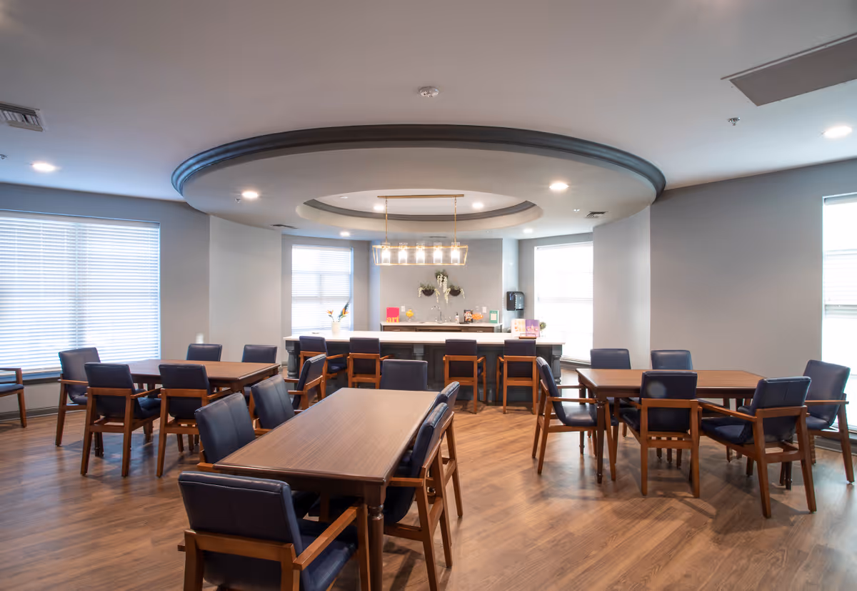 A bright dining room with multiple wooden tables and blue cushioned chairs arranged neatly on a wooden floor. The room features large windows with blinds, a modern ceiling light fixture, and a counter area with bar stools and decorative items.
