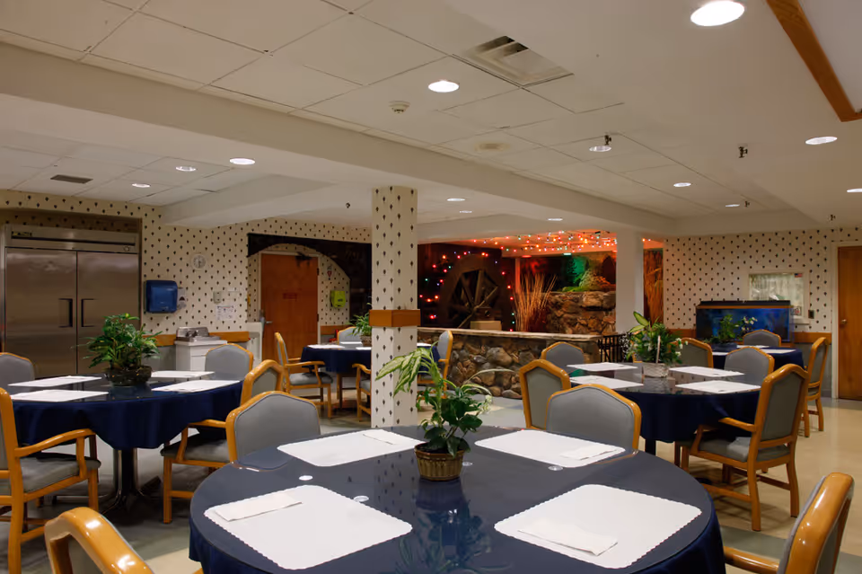 Interior view of a dining room in Marian Manor with round tables covered in dark blue tablecloths, each set with placemats and napkins. The room features wooden chairs with gray cushions, potted plants on tables, patterned wallpaper, a stone water feature with a water wheel decorated with colorful lights, and a fish tank in the background.