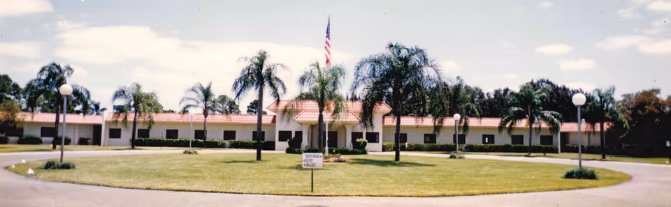 Front exterior view of a single-story rehabilitation and healthcare facility with a red-tiled roof, surrounded by palm trees and a circular driveway with a grassy center featuring an American flag on a flagpole.