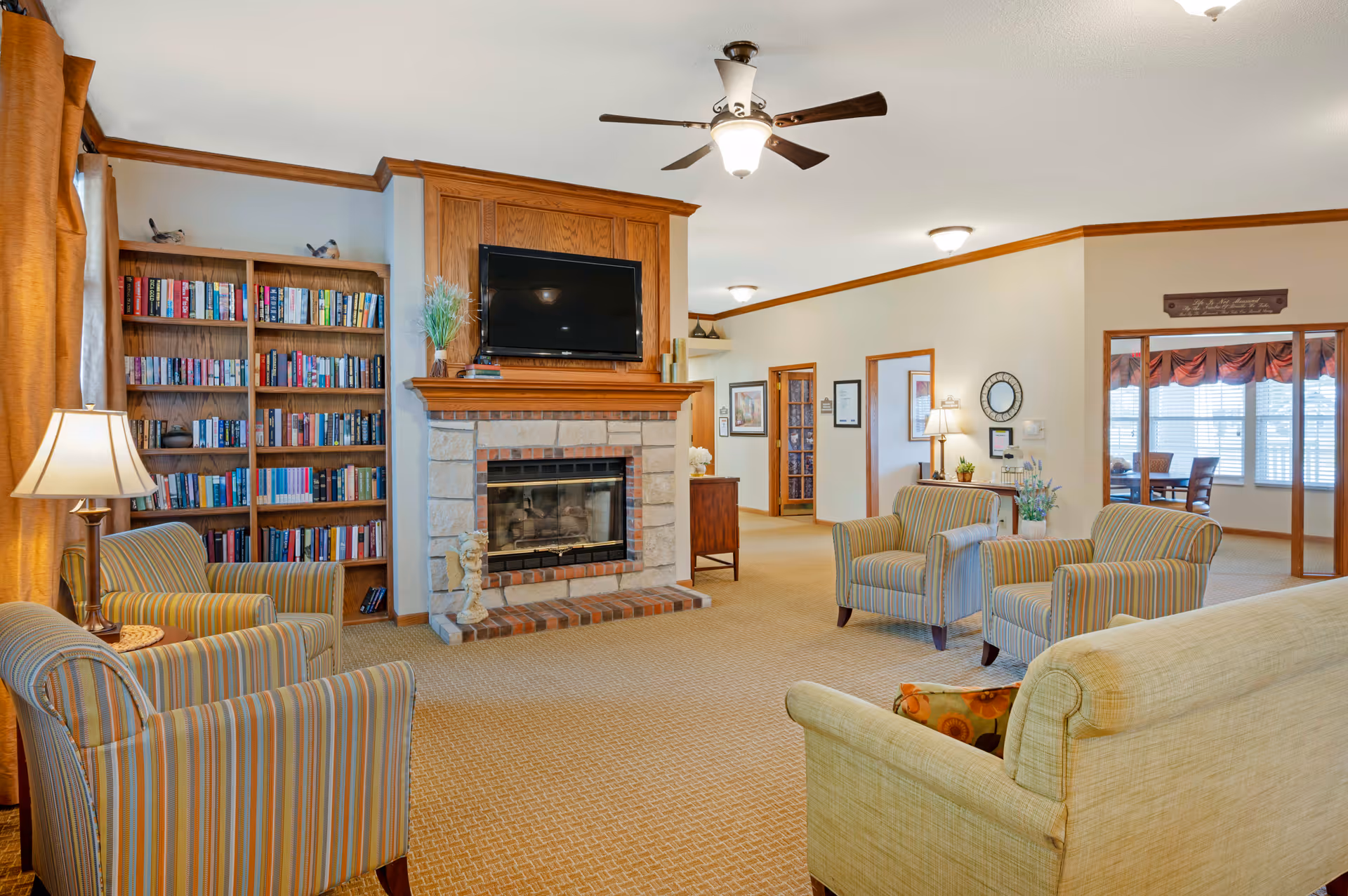 A cozy living room area in a senior living facility featuring a stone fireplace with a mounted flat-screen TV above it. There are several striped armchairs and a beige sofa arranged around the fireplace. A wooden bookshelf filled with books is positioned to the left of the fireplace. The room has warm lighting from lamps and ceiling fixtures, with beige carpeting and light-colored walls accented by wood trim. In the background, there is a dining area visible through an open doorway.