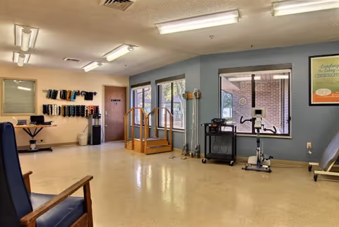 A spacious rehabilitation or physical therapy room with exercise equipment including parallel bars, a stationary bike, and weights mounted on the wall. The room has large windows letting in natural light, a chair in the foreground, and a door labeled 'Restroom'.