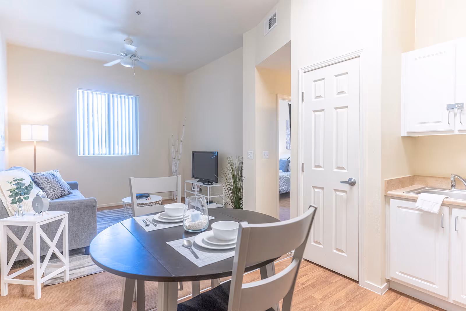 Bright open living and dining area with a round table set for two, a sofa, TV, and a small kitchenette.