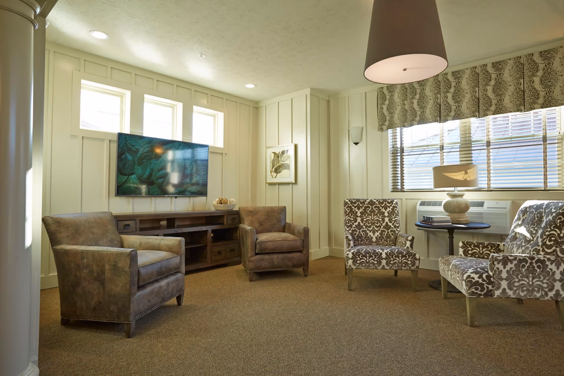 A cozy living room with four armchairs arranged around a small table with a lamp. The room has beige walls with vertical paneling, three small windows above a mounted flat-screen TV, and a larger window with blinds and a patterned valance. The floor is carpeted in a neutral tone.