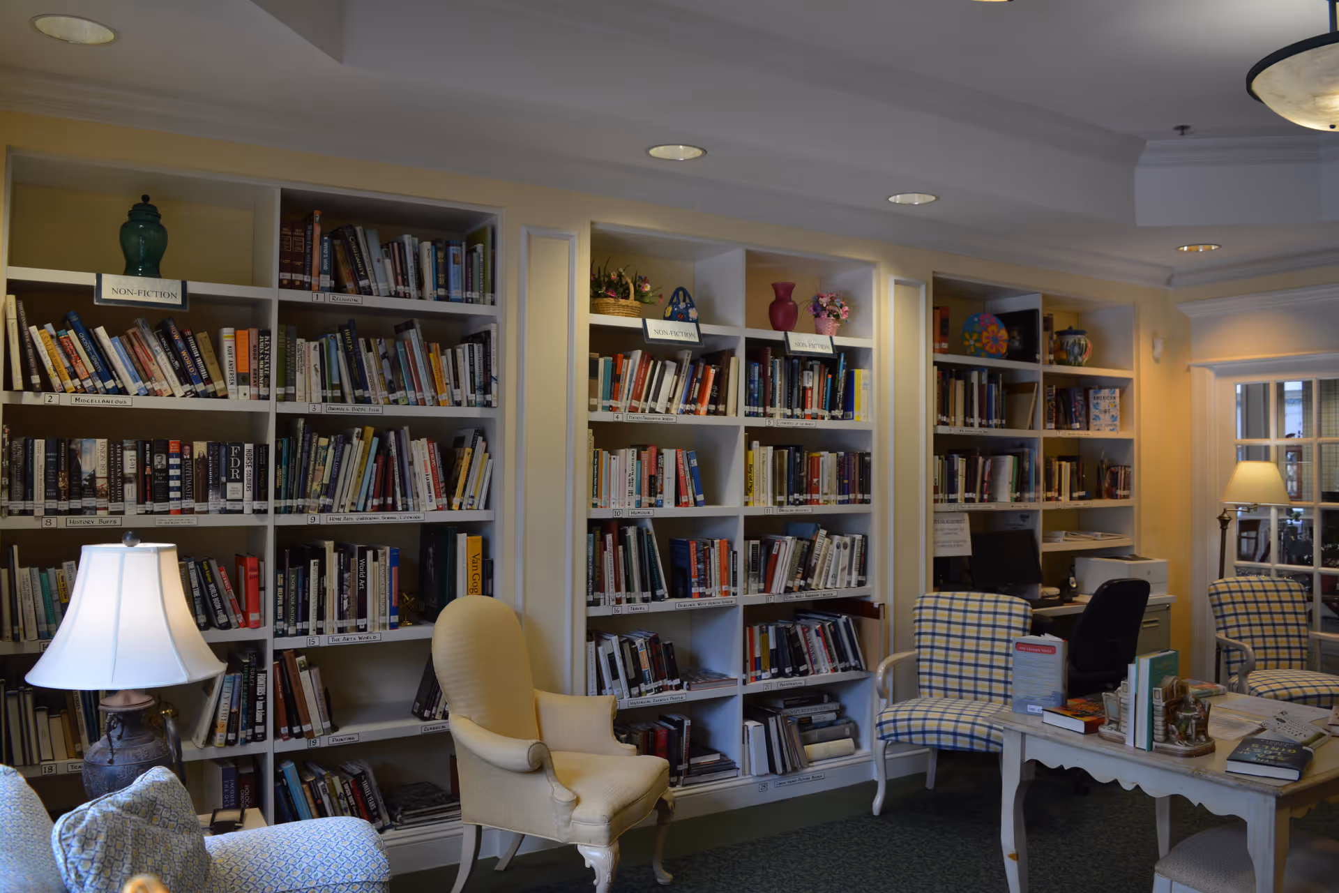 A cozy library room in The Glenwood Community featuring multiple bookshelves filled with books, comfortable armchairs, a table with books and magazines, a desk with a computer, and warm lighting from table lamps.