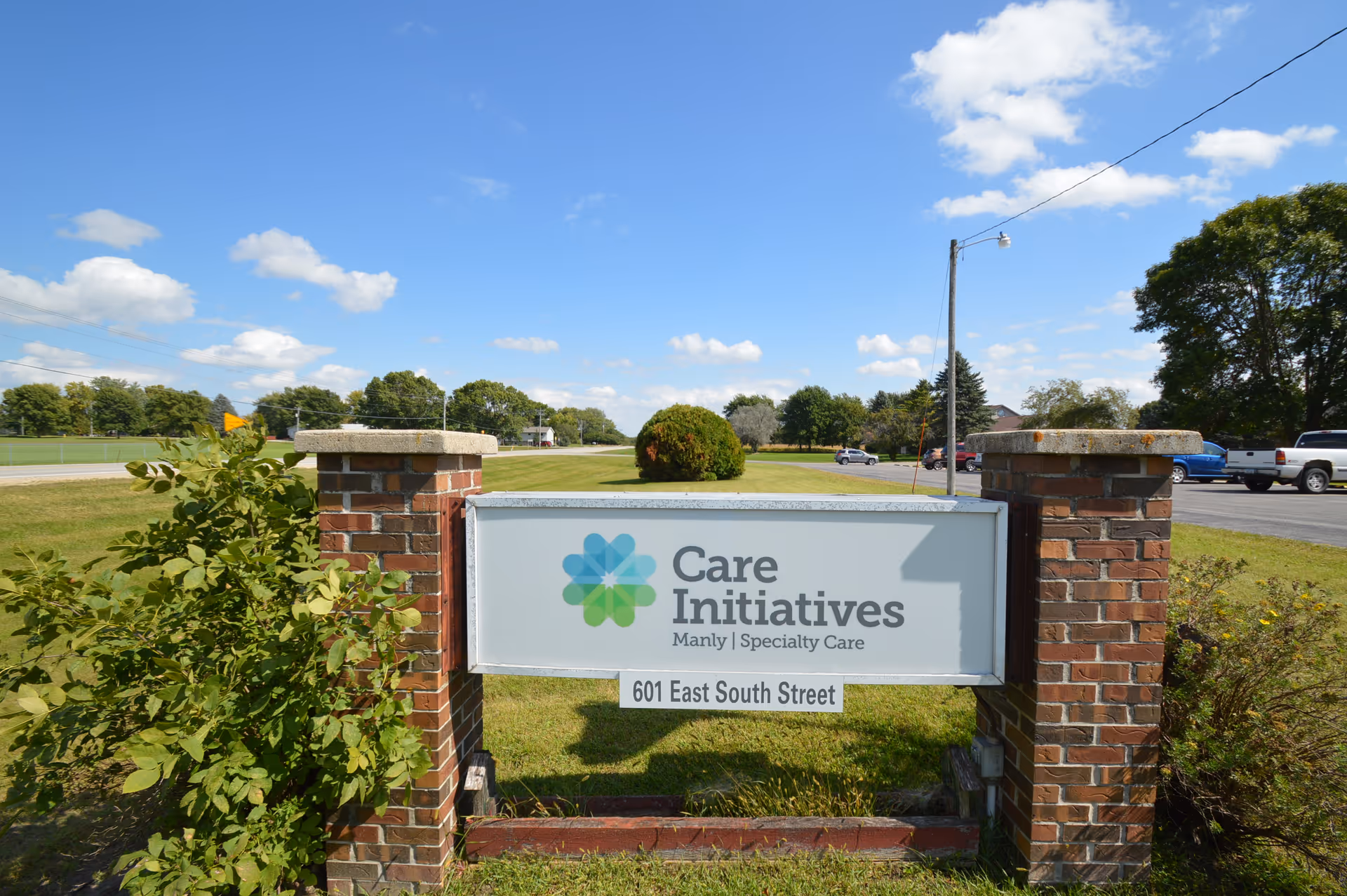 A roadside sign for Care Initiatives (Manly Specialty Care) mounted between two brick pillars on a grassy lawn under a blue sky.