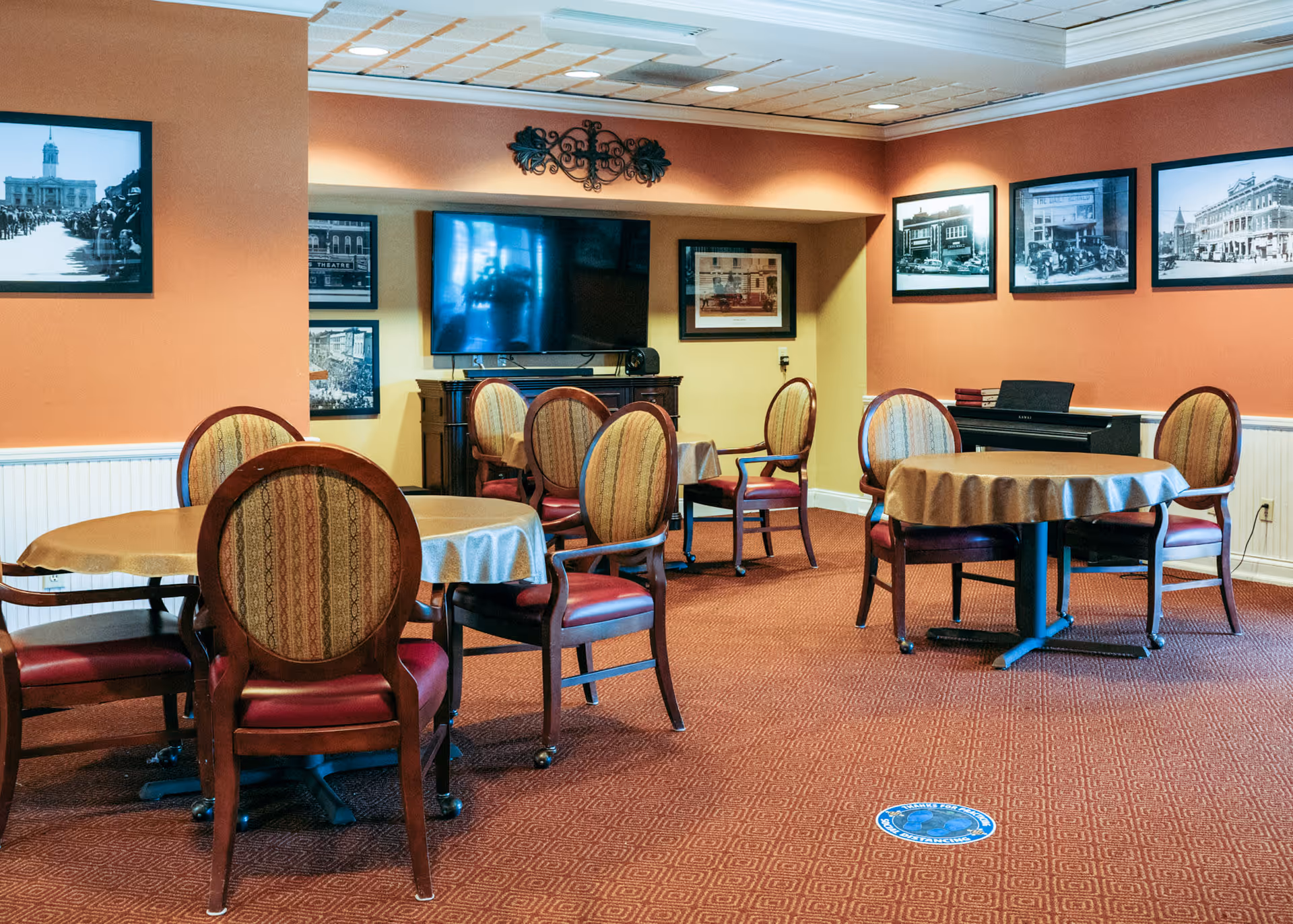A cozy common area with round tables covered with beige tablecloths and wooden chairs with patterned upholstery. The walls are painted in warm tones with framed black and white historical photographs. A flat-screen TV is mounted on the wall above a wooden cabinet, and a piano is visible in the corner. The carpet is patterned in a reddish-brown color, and there is a social distancing floor sticker visible on the carpet.