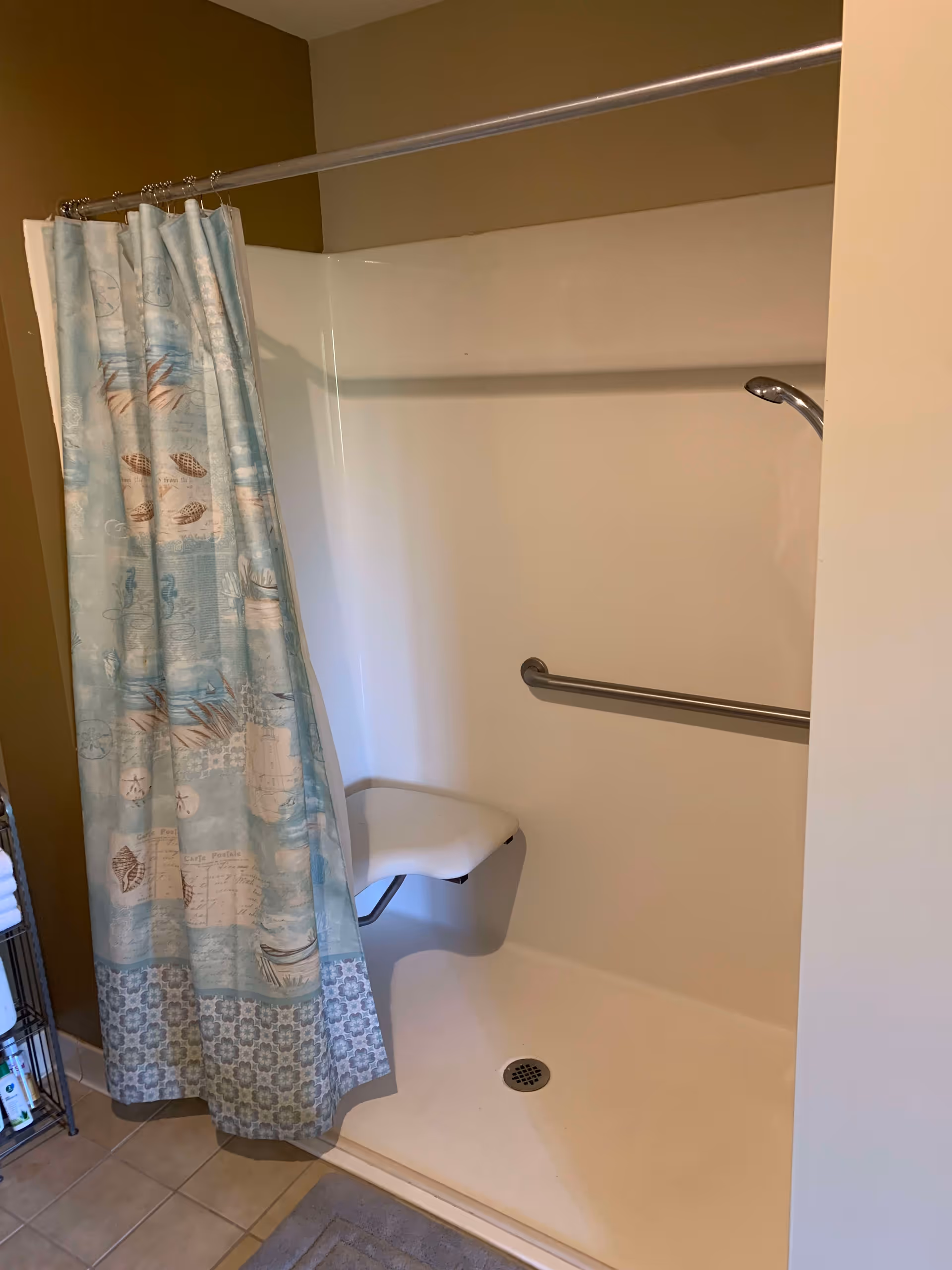 A walk-in shower with a light blue patterned shower curtain partially closed. Inside the shower, there is a white fold-down seat and a horizontal grab bar on the wall. The shower floor is beige and there is a drain in the center. The surrounding walls are light-colored, and the floor outside the shower is tiled.