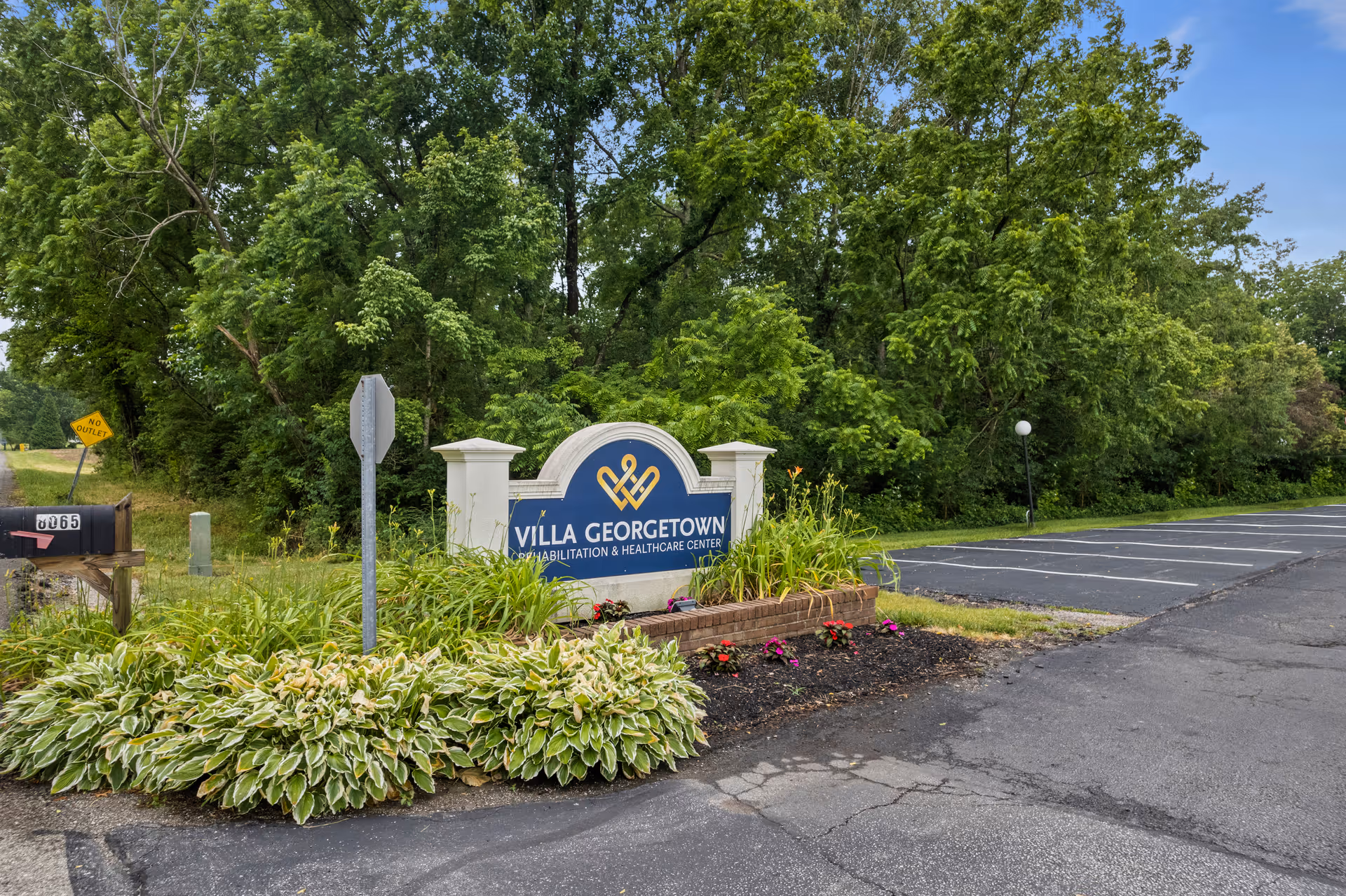 Entrance sign for Villa Georgetown Rehabilitation & Healthcare Center surrounded by greenery and plants, with a parking lot and trees in the background.