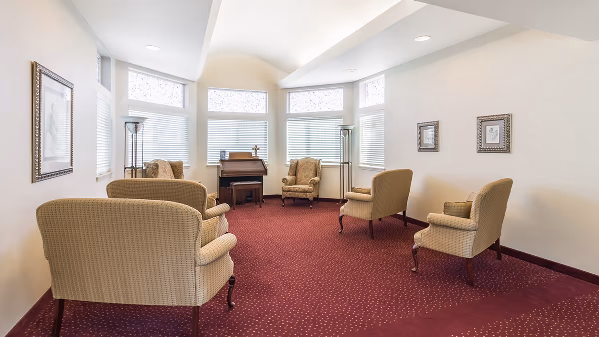 A bright and cozy sitting room with several upholstered armchairs arranged in a semi-circle on a red carpet with small yellow dots. The room has multiple windows with white blinds and decorative frosted glass panels above. There are two tall floor lamps and framed artwork on the walls. A small wooden organ with a matching stool is positioned near the windows.