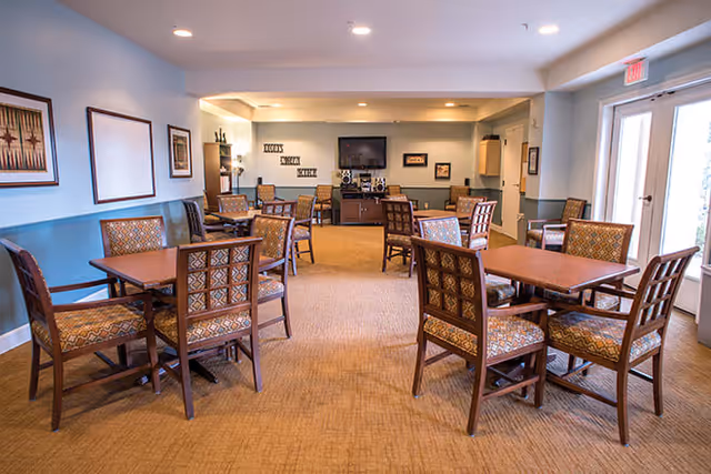 A well-lit common area with multiple wooden tables and cushioned chairs arranged for seating. The room has light blue walls with framed artwork, a carpeted floor, and a TV mounted on the far wall above a media console. There are large windows and a glass door letting in natural light.