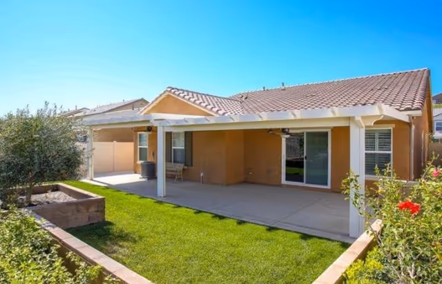 Backyard view of a single-story house with a covered patio area, green lawn, and some plants and flowers along the edges under a clear blue sky.