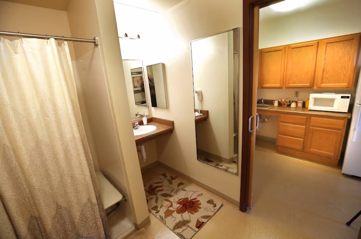 Bathroom with a shower and curtain, vanity sink and mirror, opening into a kitchenette with wooden cabinets.