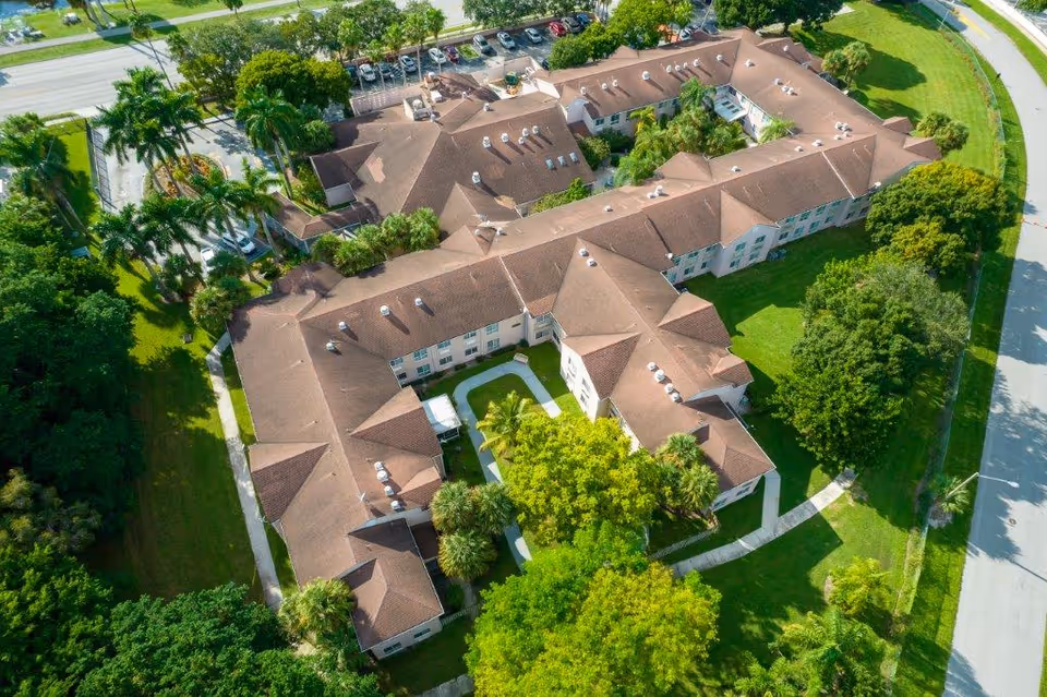 Aerial view of Colonial Assisted Living at Palm Beach showing a large, U-shaped building with a brown roof surrounded by green lawns, trees, and palm trees. There is a parking lot with cars visible at the top of the image and roads bordering the property.