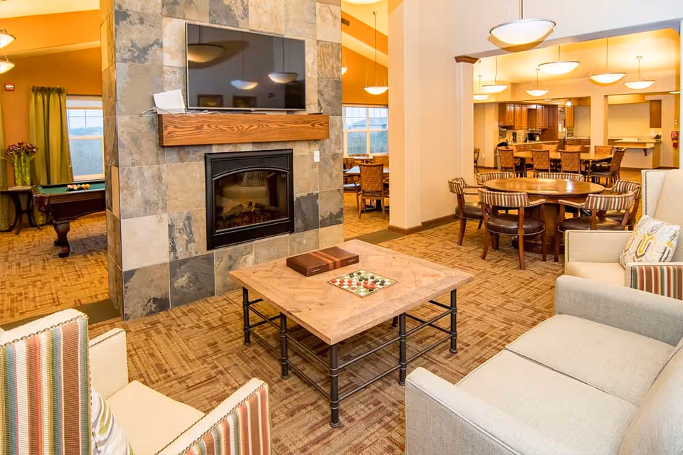 Communal lounge with sofas around a coffee table, a stone fireplace with a mounted TV, and dining and game areas in the background.