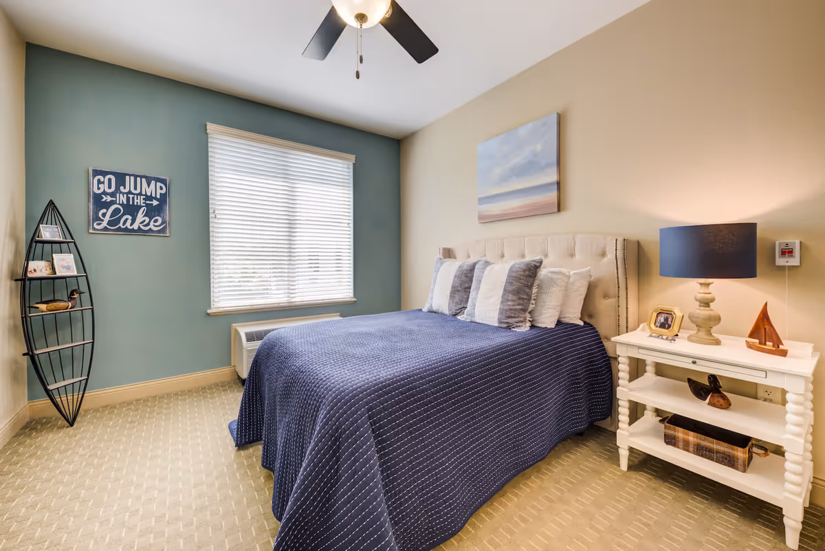A cozy bedroom with a bed covered in a navy blue quilt and several pillows. There is a white nightstand with a lamp, framed photo, and decorative items. A window with white blinds lets in natural light. The walls are painted beige and teal, with a painting above the bed and a decorative shelf shaped like a boat holding small items. A ceiling fan is visible above.