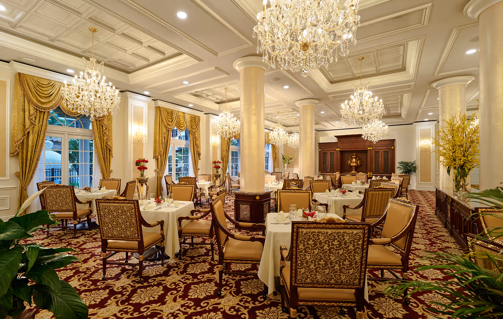 Elegant dining room with ornate chandeliers, gold draped curtains, and tables set with white tablecloths and floral centerpieces. The room features patterned carpet, large columns, and classic wooden chairs with intricate upholstery.