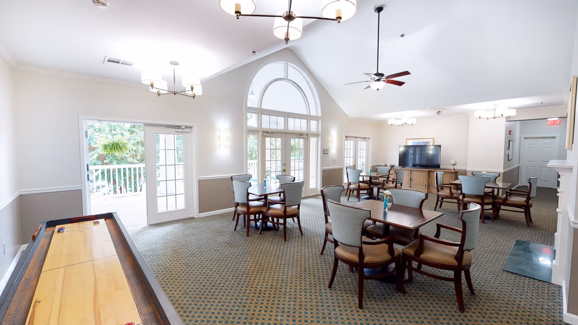 A bright and spacious common area with multiple tables and chairs arranged for seating. The room features large arched windows and glass doors letting in natural light, a ceiling fan, and modern light fixtures. There is a shuffleboard table on the left side and a large flat-screen TV on a wooden cabinet against the far wall.