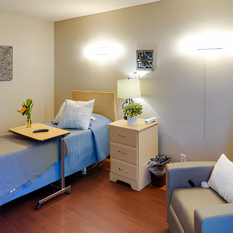 A neatly arranged bedroom in a senior living facility with a single bed covered in blue bedding, a wooden bedside table with a lamp and a small potted plant, a small overbed table with a vase of flowers and a remote control, and a gray armchair with a white pillow. The room has wooden flooring and soft lighting from wall-mounted fixtures.