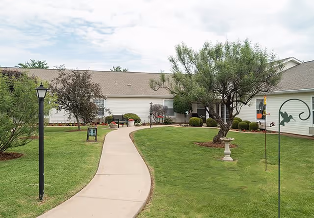 A paved walkway curves through a well-maintained green lawn with trees, shrubs, and garden decorations leading to a single-story building with white siding and a gray roof under a partly cloudy sky.