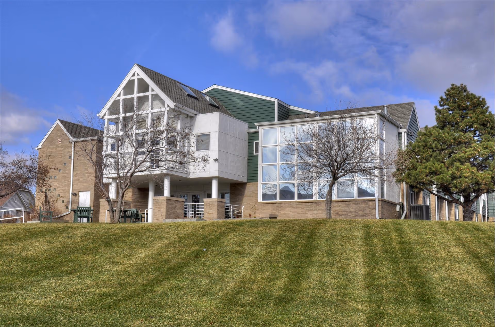 Exterior view of a multi-story assisted living facility building with large windows, a green lawn in the foreground, and a partly cloudy blue sky.