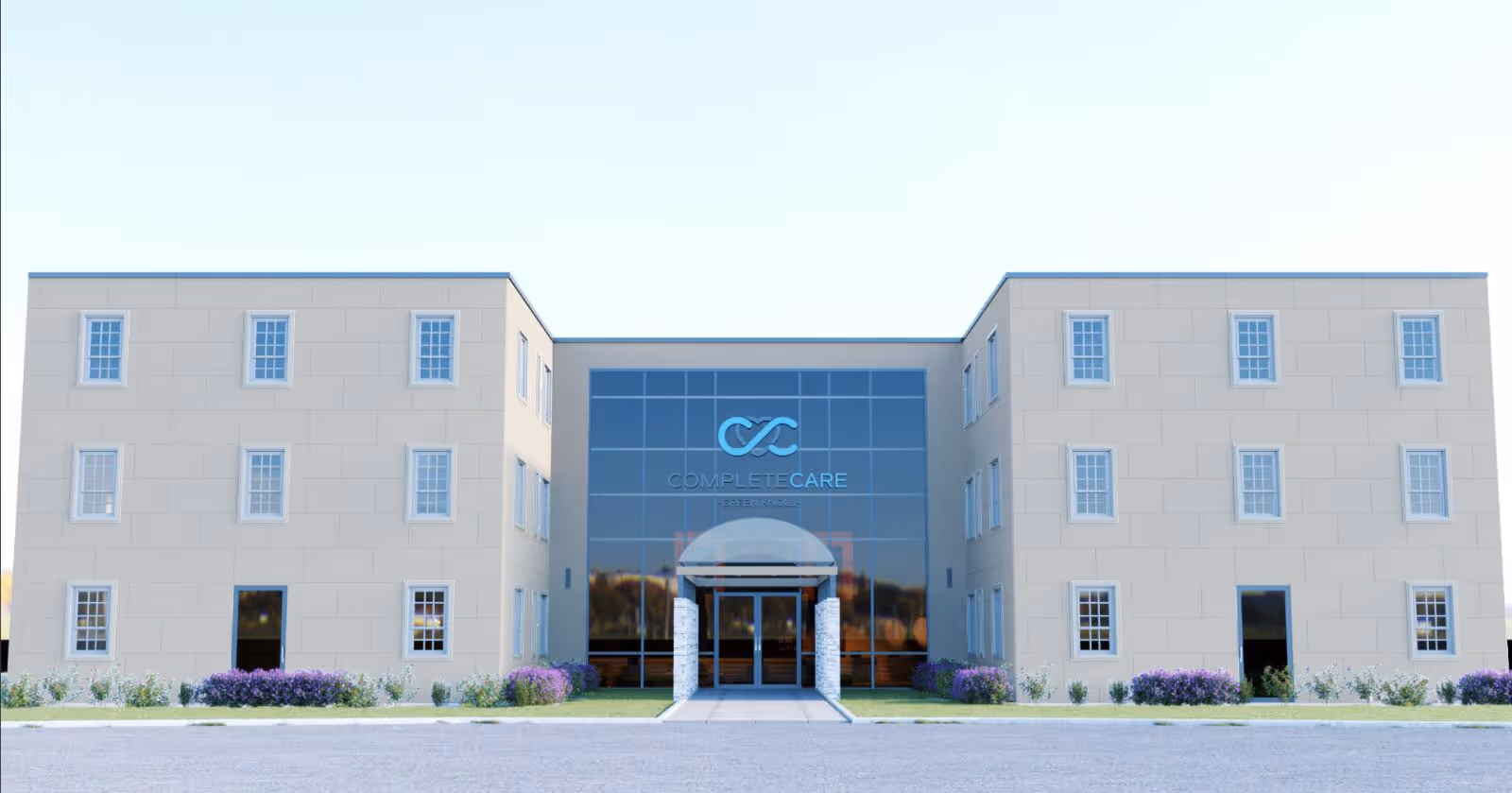 Front exterior view of a modern three-story building with beige walls and multiple windows. The center section features large glass panels with the Complete Care logo and name above the entrance. There are small bushes and purple flowers planted along the front of the building.