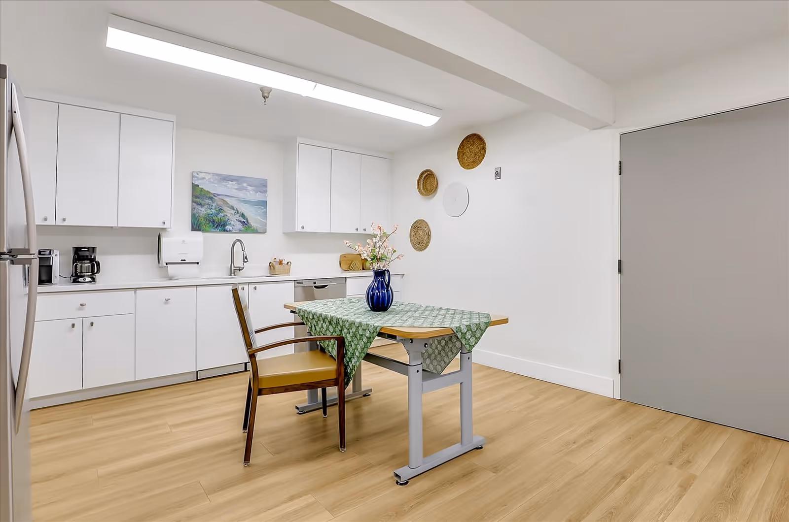 A clean and modern kitchen area with white cabinets, a stainless steel refrigerator, a coffee maker, and a dishwasher. A wooden table with a green patterned tablecloth and a blue vase with flowers is placed in the center, accompanied by a wooden chair. The walls are white with decorative woven baskets and a landscape painting. The floor is light wood, and there is a gray door on the right side.