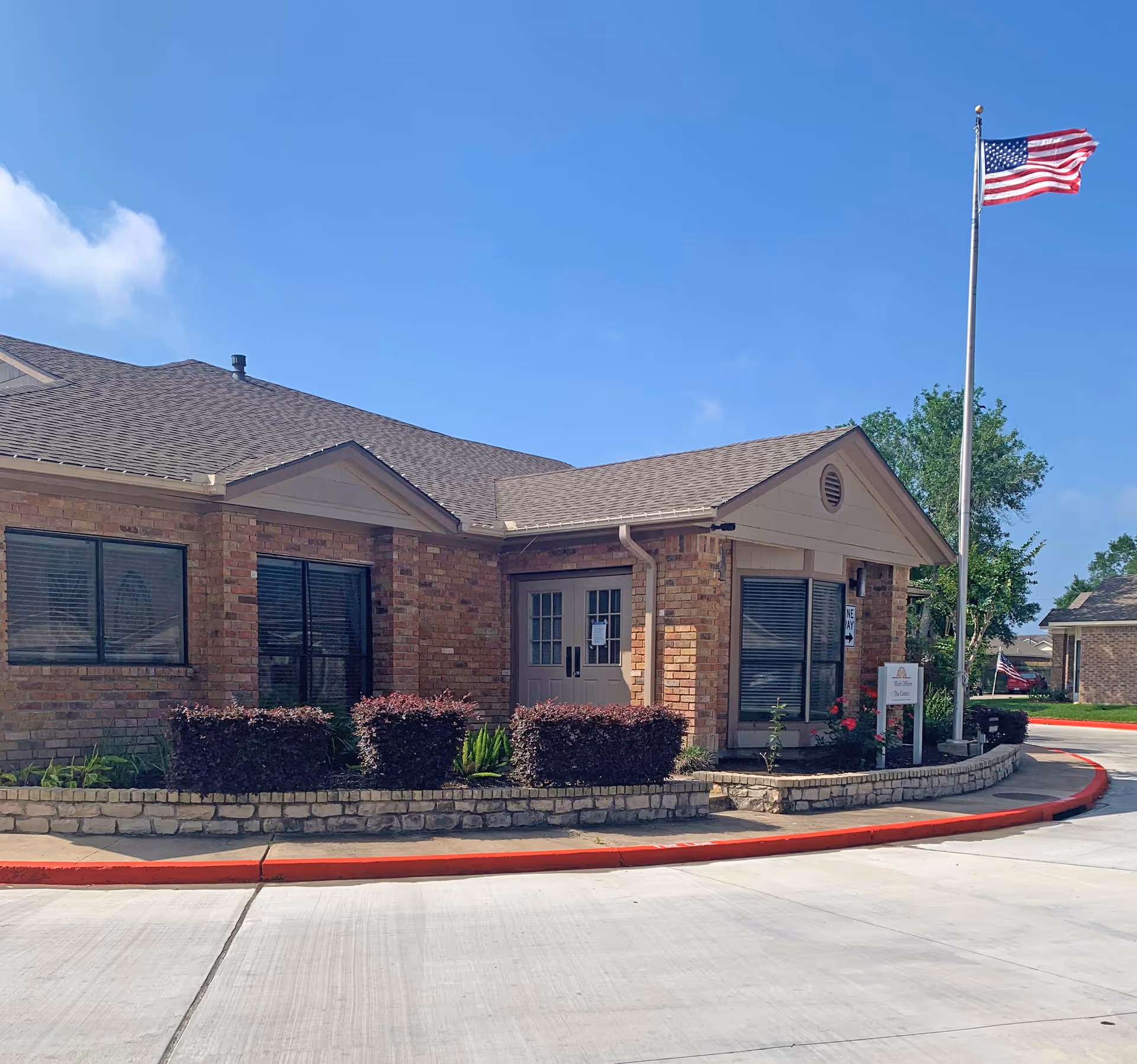 Single-story brick senior living facility entrance with an American flag on a tall pole and landscaped shrubs along a curved driveway.