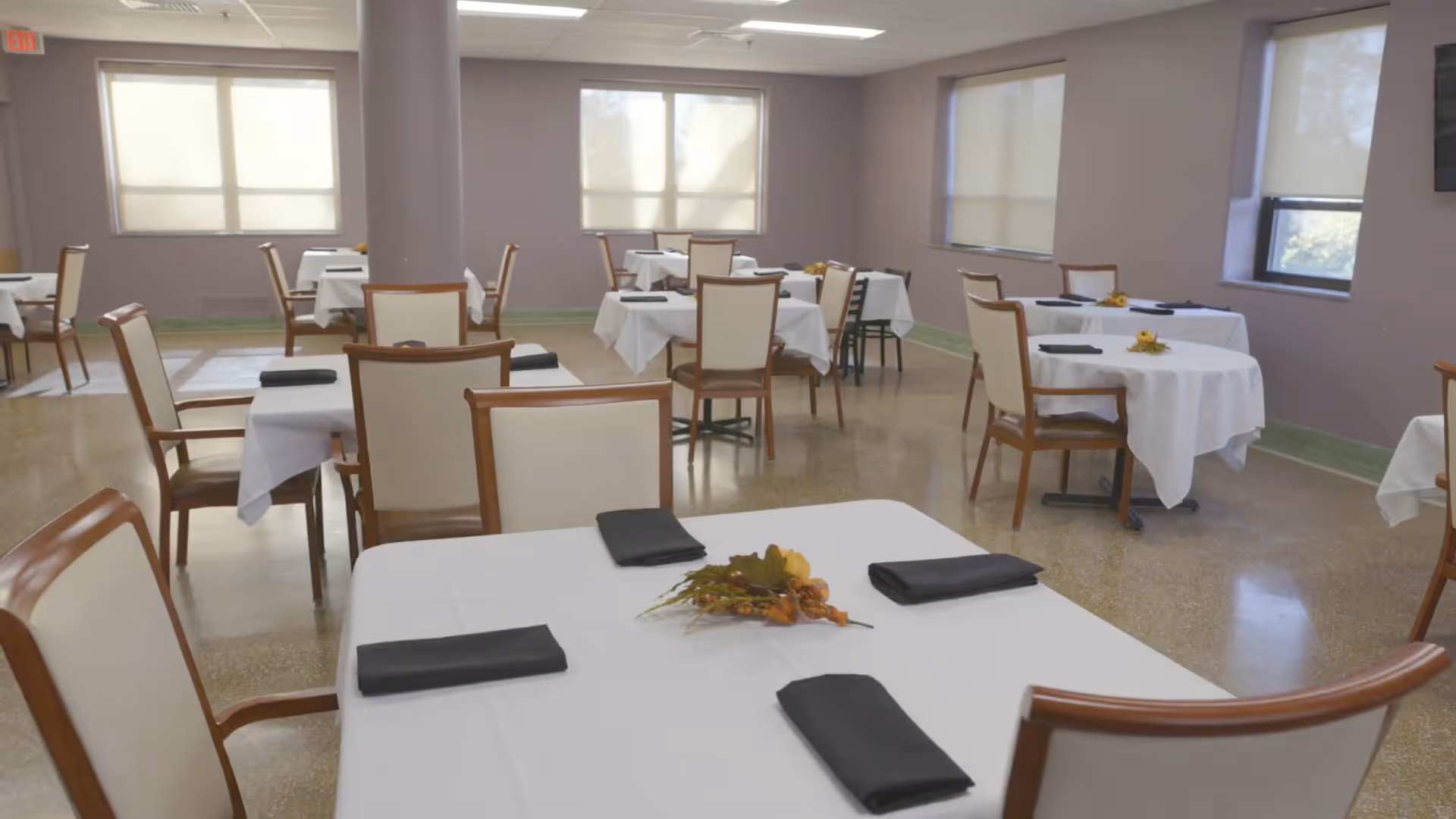 A dining room with multiple tables covered in white tablecloths, each set with black folded napkins and small floral centerpieces. The room has beige walls, several windows with blinds, and wooden chairs with cream-colored cushions.